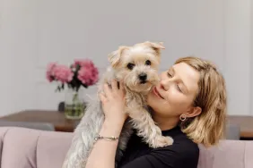 Une jeune femme embrasse un Yorkshire Terrier - une race de chien petite et câline - alors qu'elle est assise sur un canapé rose dans un salon moderne et confortable.